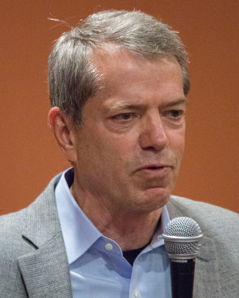 A photo of Jim Pillen from the shoulders up, an angry white man. He appears to be in his 50s or 60s, has gray hair. He is holding a microphone.