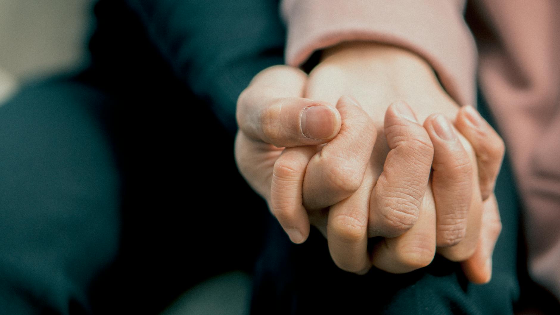 A photo of two hands, belonging to two different people. The hands are clasping each other. You can't see the people, but one person is wearing a pink sweater, the other is in dark clothes.