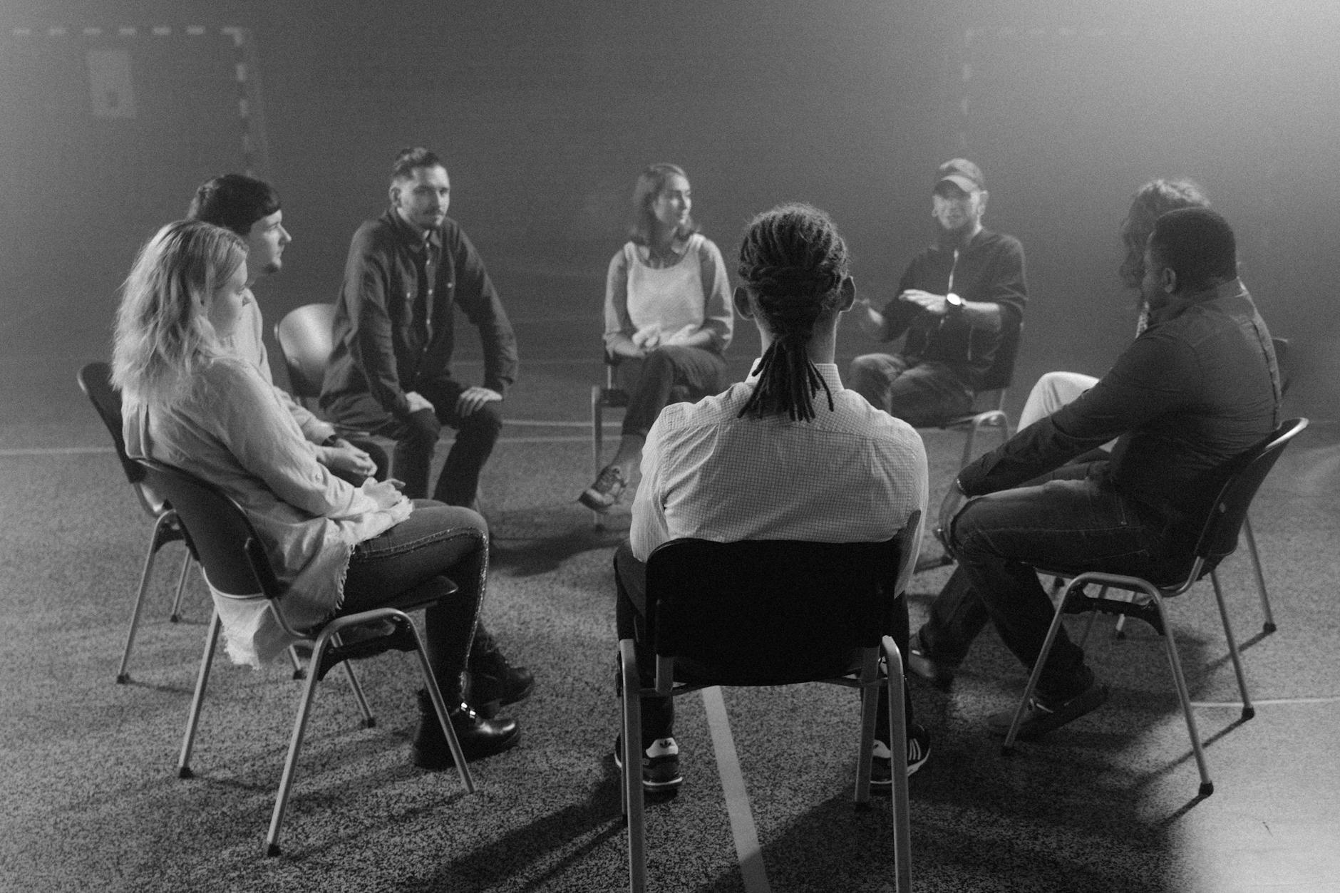 A group of people sitting in a large room in a circle, talking. It looks like some sort of support group.